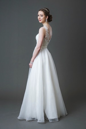 A bride wearing a long white lace and tulle gown with a fitted bodice and a flowing skirt. Her hair is styled elegantly, and she is accessorizing with large earrings and a bracelet. The background is a solid gray tone.