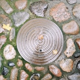 A circular manhole cover is set in a cobblestone ground, surrounded by wet stones and patches of green moss. Fallen autumn leaves are scattered on the surface, with one leaf resting on the cover itself. The metal cover features concentric circular ridges and the number 40 is visible.