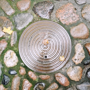 A circular manhole cover is set in a cobblestone ground, surrounded by wet stones and patches of green moss. Fallen autumn leaves are scattered on the surface, with one leaf resting on the cover itself. The metal cover features concentric circular ridges and the number 40 is visible.