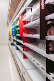 Empty shelves in a supermarket, with various sections labeled for different types of meat including beef, lamb, and sausages. The shelves are well-lit but completely bare, with no products visible.