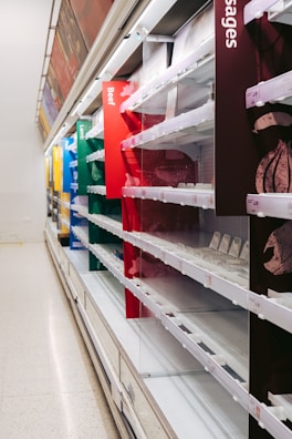 Empty shelves in a supermarket, with various sections labeled for different types of meat including beef, lamb, and sausages. The shelves are well-lit but completely bare, with no products visible.