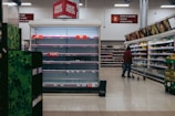An aisle in a supermarket with mostly empty shelves labeled with price tags, indicating low stock or high demand. A person with a shopping cart stands in front of a well-stocked refrigerated section containing various groceries. Overhead signs display categories such as butter, cheese, and drinks.