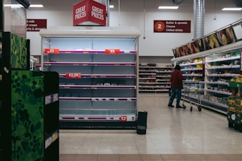 An aisle in a supermarket with mostly empty shelves labeled with price tags, indicating low stock or high demand. A person with a shopping cart stands in front of a well-stocked refrigerated section containing various groceries. Overhead signs display categories such as butter, cheese, and drinks.