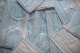 A scattered arrangement of blue disposable surgical masks, showing their rectangular shape and pleated texture. The masks are overlapping each other, creating a layered effect.