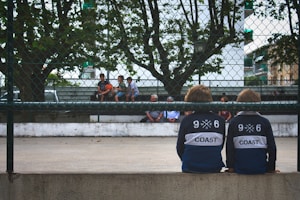 Two children wearing matching navy sweatshirts with numbers and the word 'Coast' printed on the back are sitting on a ledge. In the background, other people are seated on benches, with some holding a basketball. A chain-link fence and trees surround the area, giving a sense of a sports court or playground setting.