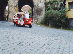 A red motorized rickshaw is driving along a cobblestone street, surrounded by historic stone arches and lush green foliage. The area exudes an old-world charm, with a person seen walking in the background.