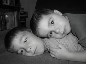 Two young children are lying down with their heads close together, displaying a calm and relaxed demeanor. They are on a textured surface, possibly a sofa, with a background that includes books and CDs on shelves.