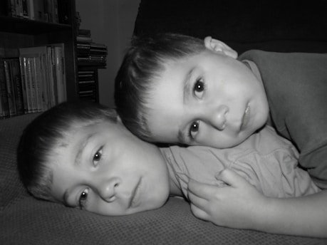 Two young children are lying down with their heads close together, displaying a calm and relaxed demeanor. They are on a textured surface, possibly a sofa, with a background that includes books and CDs on shelves.