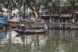 A serene riverside scene featuring traditional boats and two people wearing conical hats rowing on a wooden boat. The backdrop includes palm trees, various colorful boats docked along the waterfront, and a bustling street with motorbikes and shops.