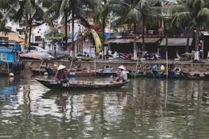 A serene riverside scene featuring traditional boats and two people wearing conical hats rowing on a wooden boat. The backdrop includes palm trees, various colorful boats docked along the waterfront, and a bustling street with motorbikes and shops.