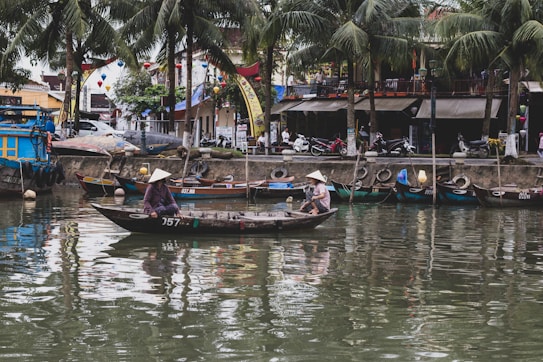A serene riverside scene featuring traditional boats and two people wearing conical hats rowing on a wooden boat. The backdrop includes palm trees, various colorful boats docked along the waterfront, and a bustling street with motorbikes and shops.