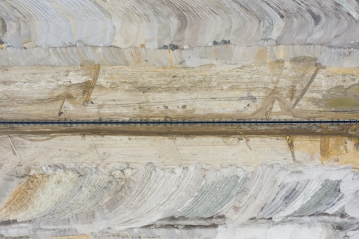 An aerial view of a mining site with layered earth tones and a long, dark road or conveyor belt running horizontally across the center. Earth-moving equipment is visible near the bottom right, amidst large, sweeping patterns in the terrain that suggest excavation or quarrying activity.