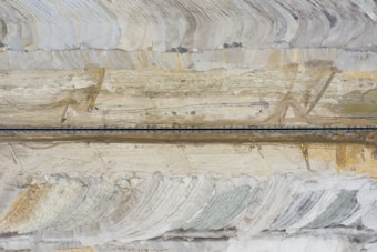 An aerial view of a mining site with layered earth tones and a long, dark road or conveyor belt running horizontally across the center. Earth-moving equipment is visible near the bottom right, amidst large, sweeping patterns in the terrain that suggest excavation or quarrying activity.