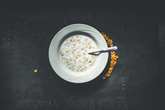 A white bowl filled with milk and cereal placed on a dark background. A spoon rests inside the bowl, and some cereal pieces are scattered around the bowl. The lighting creates a subtle shadow effect, giving a moody ambiance.