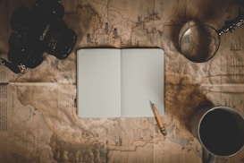 A top-down view of an arrangement featuring a vintage camera, an open blank notebook with a pencil on top, a map underneath, a magnifying glass, and a cup of coffee. The items suggest a theme of travel or exploration with an emphasis on planning or documenting adventures.