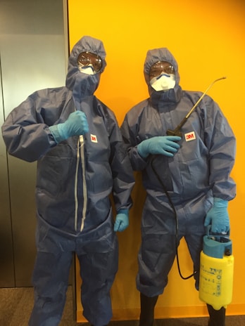 Two individuals dressed in full protective gear, including blue hazmat suits, gloves, masks, and goggles. One person is holding a sprayer, and both have 3M logos on their uniforms. They are standing against a bright yellow wall.