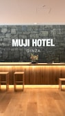 A hotel reception area features a rustic stone wall with the words 'MUJI HOTEL GINZA' prominently displayed. The space includes a wooden front desk with integrated lighting and a minimalist floral arrangement on top. Two simple wooden stools are placed in front of the desk, and the flooring is made of light-colored wood.