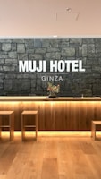A hotel reception area features a rustic stone wall with the words 'MUJI HOTEL GINZA' prominently displayed. The space includes a wooden front desk with integrated lighting and a minimalist floral arrangement on top. Two simple wooden stools are placed in front of the desk, and the flooring is made of light-colored wood.