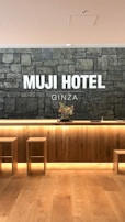 A hotel reception area features a rustic stone wall with the words 'MUJI HOTEL GINZA' prominently displayed. The space includes a wooden front desk with integrated lighting and a minimalist floral arrangement on top. Two simple wooden stools are placed in front of the desk, and the flooring is made of light-colored wood.
