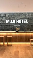 A hotel reception area features a rustic stone wall with the words 'MUJI HOTEL GINZA' prominently displayed. The space includes a wooden front desk with integrated lighting and a minimalist floral arrangement on top. Two simple wooden stools are placed in front of the desk, and the flooring is made of light-colored wood.
