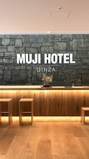A hotel reception area features a rustic stone wall with the words 'MUJI HOTEL GINZA' prominently displayed. The space includes a wooden front desk with integrated lighting and a minimalist floral arrangement on top. Two simple wooden stools are placed in front of the desk, and the flooring is made of light-colored wood.