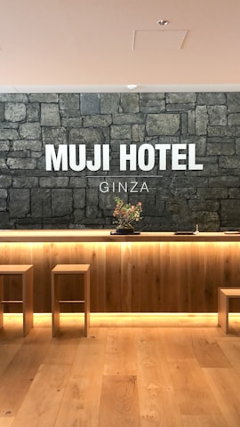 A hotel reception area features a rustic stone wall with the words 'MUJI HOTEL GINZA' prominently displayed. The space includes a wooden front desk with integrated lighting and a minimalist floral arrangement on top. Two simple wooden stools are placed in front of the desk, and the flooring is made of light-colored wood.