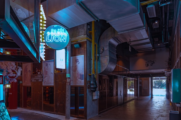 An industrial-style interior corridor with exposed ducts and pipes features vintage posters on wooden panels lining the walls. A neon sign with Japanese characters and the word 'LION' emits a blue glow. Various ambient warm lights illuminate the scene, adding a cozy ambiance.
