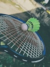 A green badminton shuttlecock rests on a tennis racket's strings in an outdoor setting. The focus is on the sporting equipment, with a blurred background that includes grass and a wall.