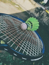 A green badminton shuttlecock rests on a tennis racket's strings in an outdoor setting. The focus is on the sporting equipment, with a blurred background that includes grass and a wall.