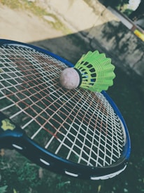 A green badminton shuttlecock rests on a tennis racket's strings in an outdoor setting. The focus is on the sporting equipment, with a blurred background that includes grass and a wall.
