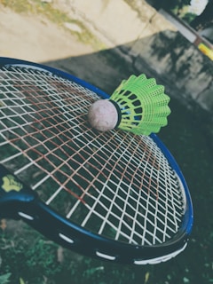 A green badminton shuttlecock rests on a tennis racket's strings in an outdoor setting. The focus is on the sporting equipment, with a blurred background that includes grass and a wall.