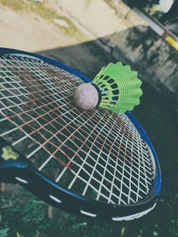 A green badminton shuttlecock rests on a tennis racket's strings in an outdoor setting. The focus is on the sporting equipment, with a blurred background that includes grass and a wall.