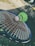 A green badminton shuttlecock rests on a tennis racket's strings in an outdoor setting. The focus is on the sporting equipment, with a blurred background that includes grass and a wall.