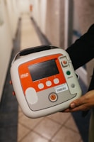 A person is holding a medical device, specifically an automated external defibrillator (AED), with a red accent and labeled 'iPAD'. The device has a screen, control buttons, and an on/off switch. The hallway setting in the background includes tiles on the floor.
