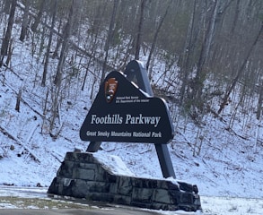 A large sign for Foothills Parkway in the Great Smoky Mountains National Park is displayed against a backdrop of snowy trees. The sign features the National Park Service emblem and is supported by a stone base.
