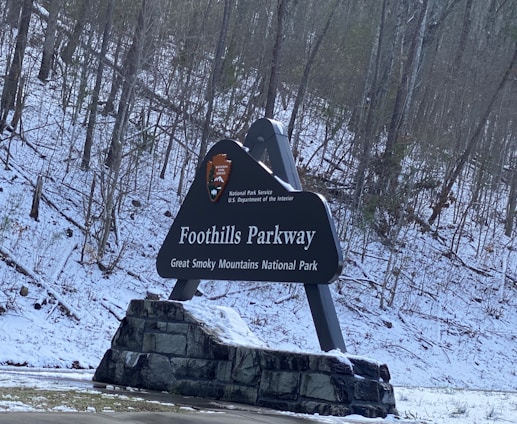 A large sign for Foothills Parkway in the Great Smoky Mountains National Park is displayed against a backdrop of snowy trees. The sign features the National Park Service emblem and is supported by a stone base.