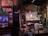 A vintage-style music venue room features a small stage with a wooden piano and a tip bucket. An old American flag hangs on the wall alongside weathered posters and framed photographs. A guitar amplifier and a stool are placed near the stage. The walls are a mix of exposed brick and peeling plaster, contributing to the rustic ambiance. Lighting is dim and atmospheric.
