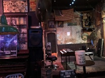 A vintage-style music venue room features a small stage with a wooden piano and a tip bucket. An old American flag hangs on the wall alongside weathered posters and framed photographs. A guitar amplifier and a stool are placed near the stage. The walls are a mix of exposed brick and peeling plaster, contributing to the rustic ambiance. Lighting is dim and atmospheric.