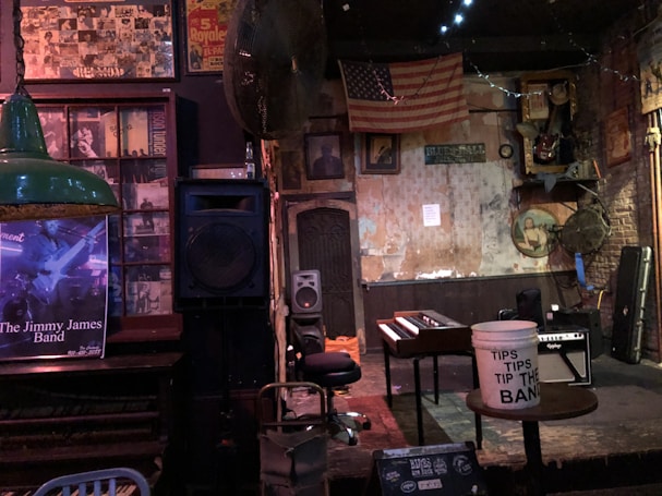A vintage-style music venue room features a small stage with a wooden piano and a tip bucket. An old American flag hangs on the wall alongside weathered posters and framed photographs. A guitar amplifier and a stool are placed near the stage. The walls are a mix of exposed brick and peeling plaster, contributing to the rustic ambiance. Lighting is dim and atmospheric.