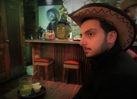 A person wearing a cowboy hat sits at a wooden table in a rustic bar setting. The bar features a fun display of colorful gumballs in a jar, and in the background, a poster of a cowboy adds to the thematic decor. The lighting is warm, contributing to a cozy ambiance.