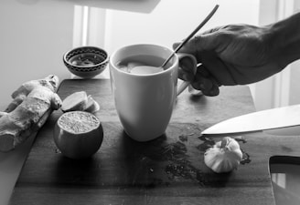 A hand is holding a white mug with a spoon inside it, placed on a wooden cutting board. Surrounding the mug are a knife, a clove of garlic, a halved lime, a piece of ginger, and a small bowl. The setting suggests preparation for a homemade remedy or drink.