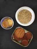 A variety of baked goods are displayed against a dark surface. There is a round white ceramic bowl containing a grainy, crumbly mixture, possibly a type of spice blend or crushed biscuits. A smaller, clear plastic container holds several light brown, round biscuits. A metal tin, also with several round biscuits, features a darker brown color and a slightly textured surface.