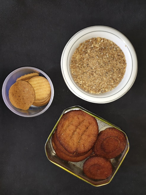 A variety of baked goods are displayed against a dark surface. There is a round white ceramic bowl containing a grainy, crumbly mixture, possibly a type of spice blend or crushed biscuits. A smaller, clear plastic container holds several light brown, round biscuits. A metal tin, also with several round biscuits, features a darker brown color and a slightly textured surface.