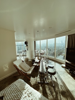 A modern living space featuring a music setup including a keyboard, drum set, and microphones. Large windows provide a panoramic city view, creating an open and airy atmosphere. The room is sparsely furnished with a lounge chair and TV, capturing abundant natural light.