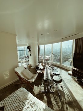 A modern living space featuring a music setup including a keyboard, drum set, and microphones. Large windows provide a panoramic city view, creating an open and airy atmosphere. The room is sparsely furnished with a lounge chair and TV, capturing abundant natural light.