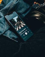 A smartphone displaying a music streaming application with an album cover visible on the screen. The phone is placed on denim fabric with a pair of black headphones partially visible alongside.