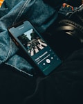 A smartphone displaying a music streaming application with an album cover visible on the screen. The phone is placed on denim fabric with a pair of black headphones partially visible alongside.