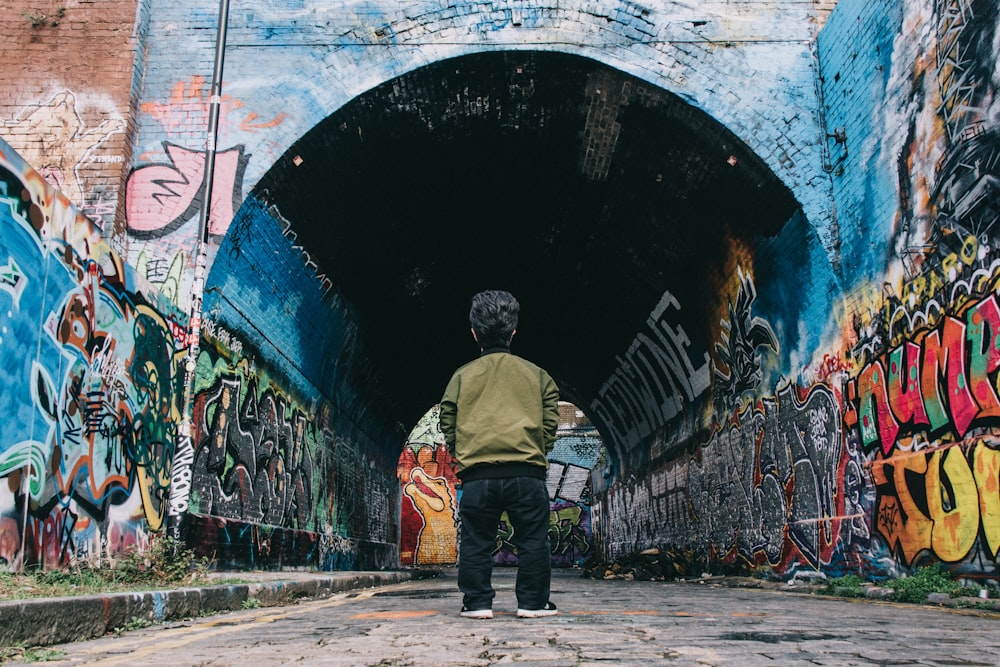 Hombre con chaqueta gris y jeans de mezclilla azul de pie junto a la pared de graffiti durante el día