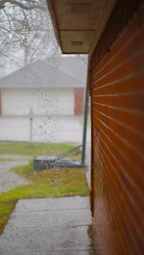 Water pours off the edge of a roof, flowing near the wall of a red brick building. The background reveals a foggy atmosphere with a leafless tree and residential homes. The visible path is wet, indicating recent rain.