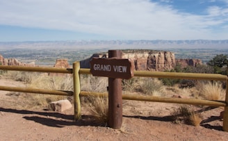 A scenic overlook featuring a sign labeled 'Grand View' situated next to a wooden fence. In the background, expansive rocky mesas and a wide valley stretch towards a distant mountain range under a partly cloudy sky.
